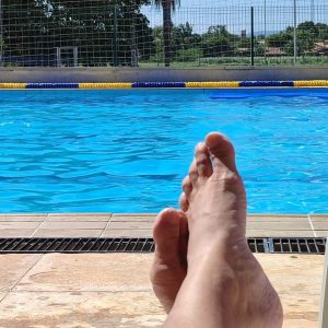 Pés descansando na borda de uma piscina azul em meio a um sol gostoso de verão